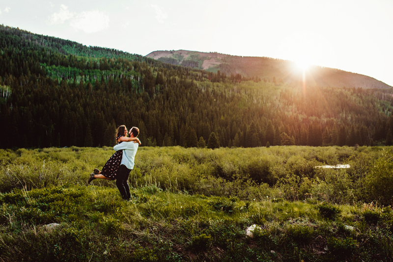 Elopement Photographer, a man embraces and lifts his partner into the air. She is smiling. they are in a lush green meadow with a forest behind them.