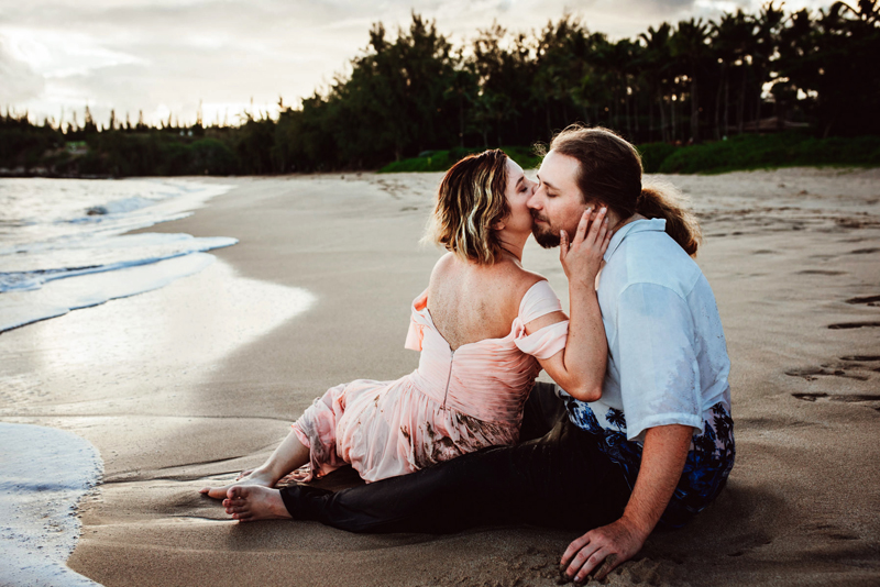 Elopement photographer, woman leans back to kiss man as they sit in the sand directly before the lakes shore, her dress wet with sand