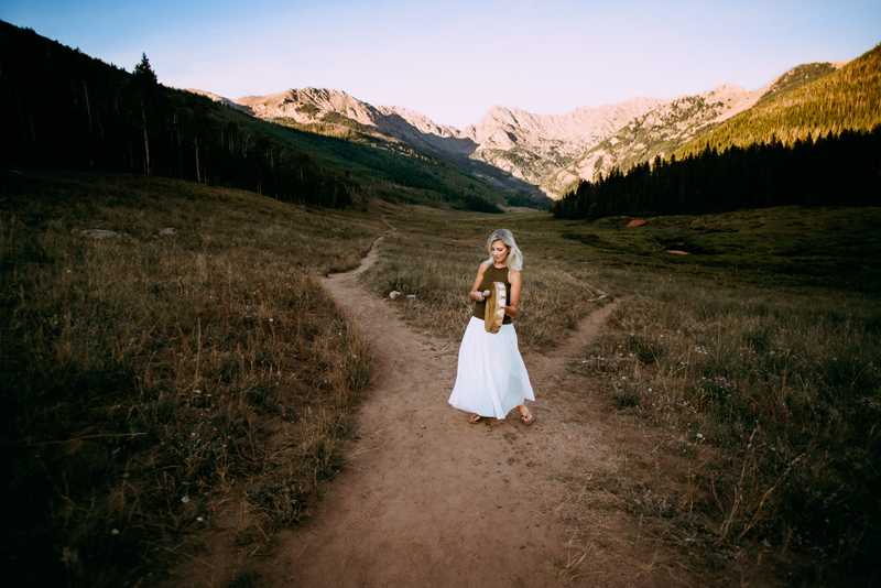 Business Photography,  Blonde Woman stands at the fork of two mountain trails. She has a sleeveless olive colored blouse and a white long skirt. She plays a unique style of drum, culturally decorated with wood inlays