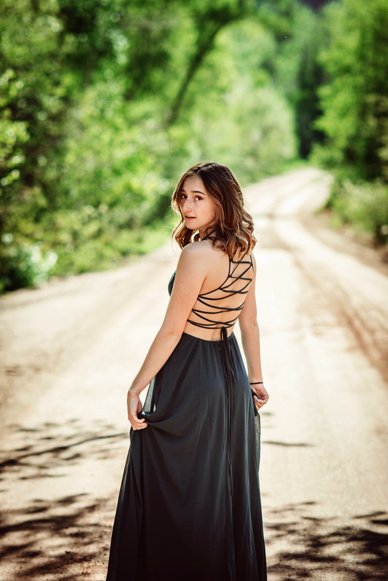 Senior Portrait, High School woman in dark dress looks back as she walks on forest road