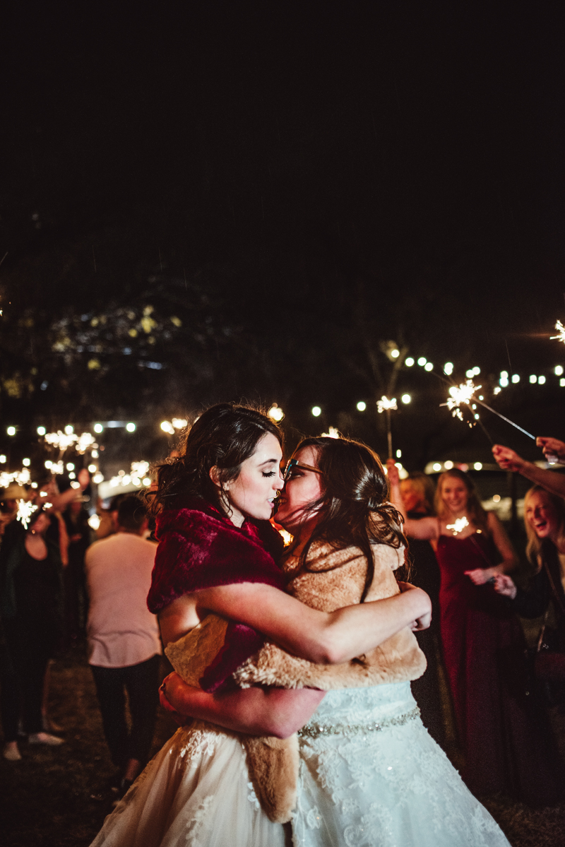Elopement photographer, two brides kiss with their arms wrapped around each other as people hold sparklers in the air