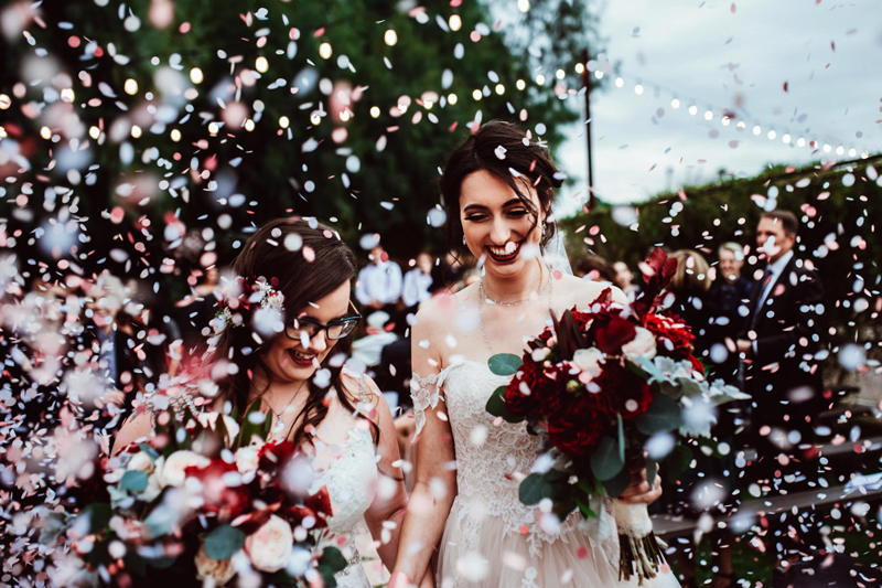 Wedding Photographer, two brides hold hands and bouquets of roses in a sea of confetti as crowds cheer