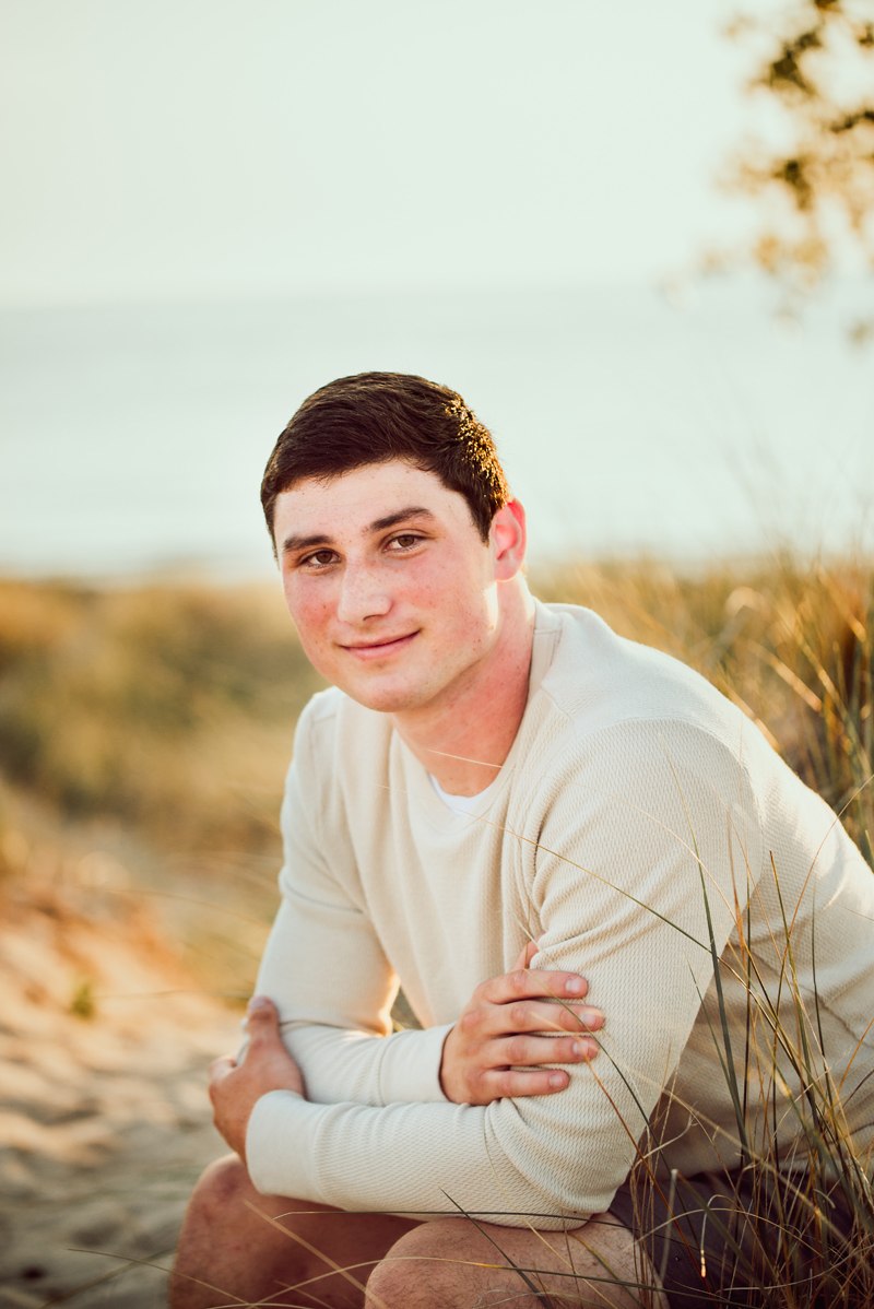 Senior Portrait, High School man in white knit sweater and shorts sits near grass and sand smiling