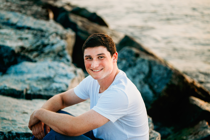 Senior Portrait, High School brunette man in white t-shirt sits on rocks near ocean