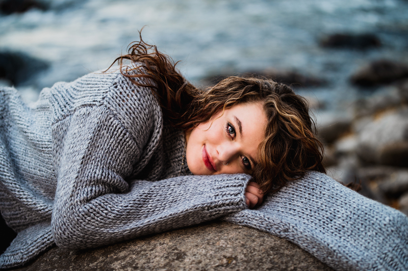 Senior Portrait, High School woman with brunette wavy hair lays on her arm on a rock near river