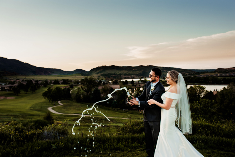 Engagement Photographer,A bride and groom stand in a lush green lakeside and pop a bottle of champagne
