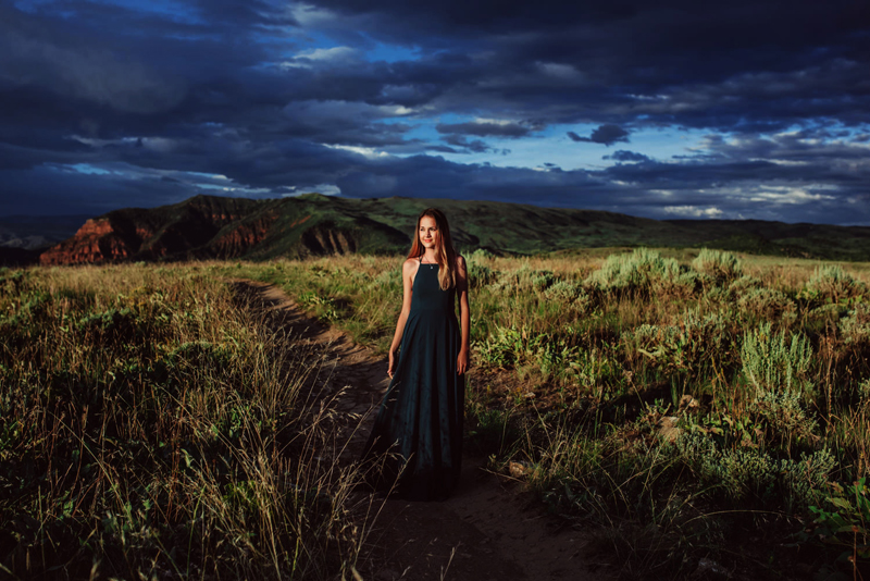 Senior Portrait, High School woman in classy sleek dress walks on grassy hillside trails at dusk