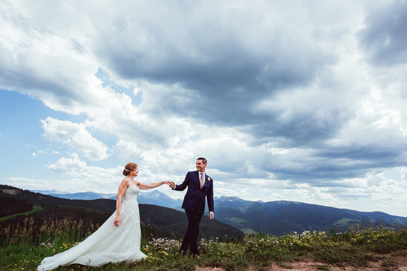 Elopement Photographer, Man leads bride in her wedding dress through flowery hillside with mountains behind them