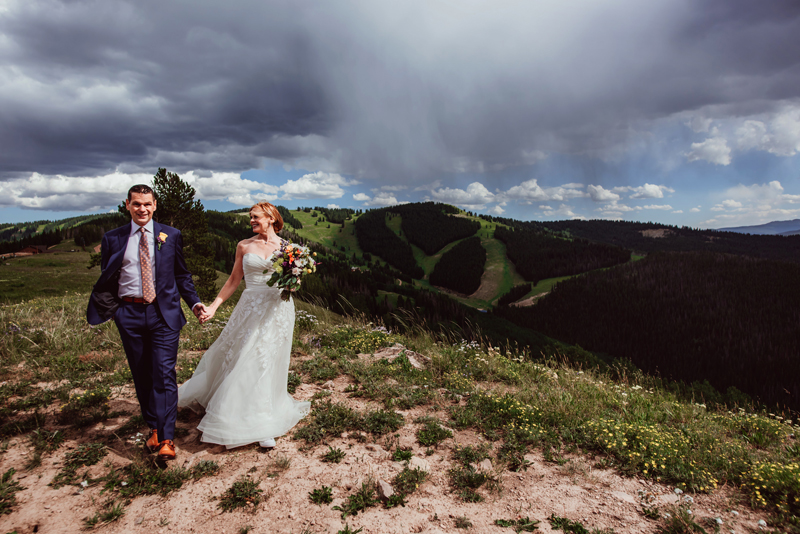 Elopement Photographer, a bride with her gown and bouquet of colorful flowers holds hands with her groom on a walk along a green hillside